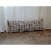A long rectangular lumbar pillow with a textured plaid pattern in beige, brown, and gray, resting against a white curtain on a concrete floor.
