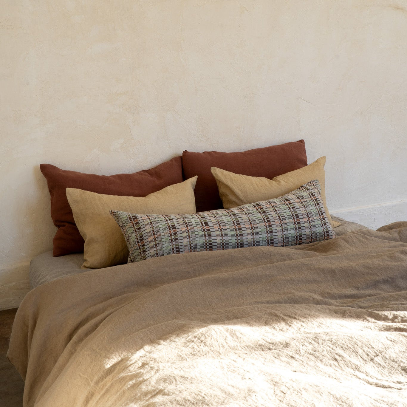 A bed styled with a textured beige duvet cover and an assortment of pillows, including two rust-brown square pillows, two tan rectangular pillows, and a plaid lumbar pillow, set against a warm, textured plaster wall.
