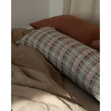 A long lumbar pillow with a geometric woven pattern in muted green, brown, and beige, resting on a bed with brown sheets and a rust-colored pillowcase.