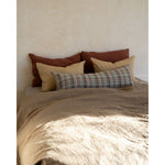A bed styled with a textured beige duvet cover and a collection of pillows, including two rust-brown pillows, two tan pillows, and a plaid lumbar pillow, set against a textured beige plaster wall with soft sunlight filtering in.