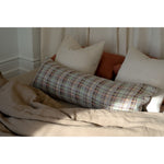 A long rectangular lumbar pillow with a woven geometric pattern in muted blue, green, beige, and brown tones, styled on a bed with neutral beige linens and other pillows.