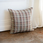 A square throw pillow with a geometric plaid pattern in beige, brown, gray, and muted green, leaning against a white wall on a textured concrete floor.