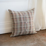 A square throw pillow with a geometric plaid pattern in muted beige, brown, gray, and soft green tones, leaning against a white paneled wall on a textured concrete floor.
