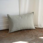 A rectangular linen throw pillow in a muted sage green color, leaning against a white wall on a textured brown floor with soft natural light filtering in from the right.