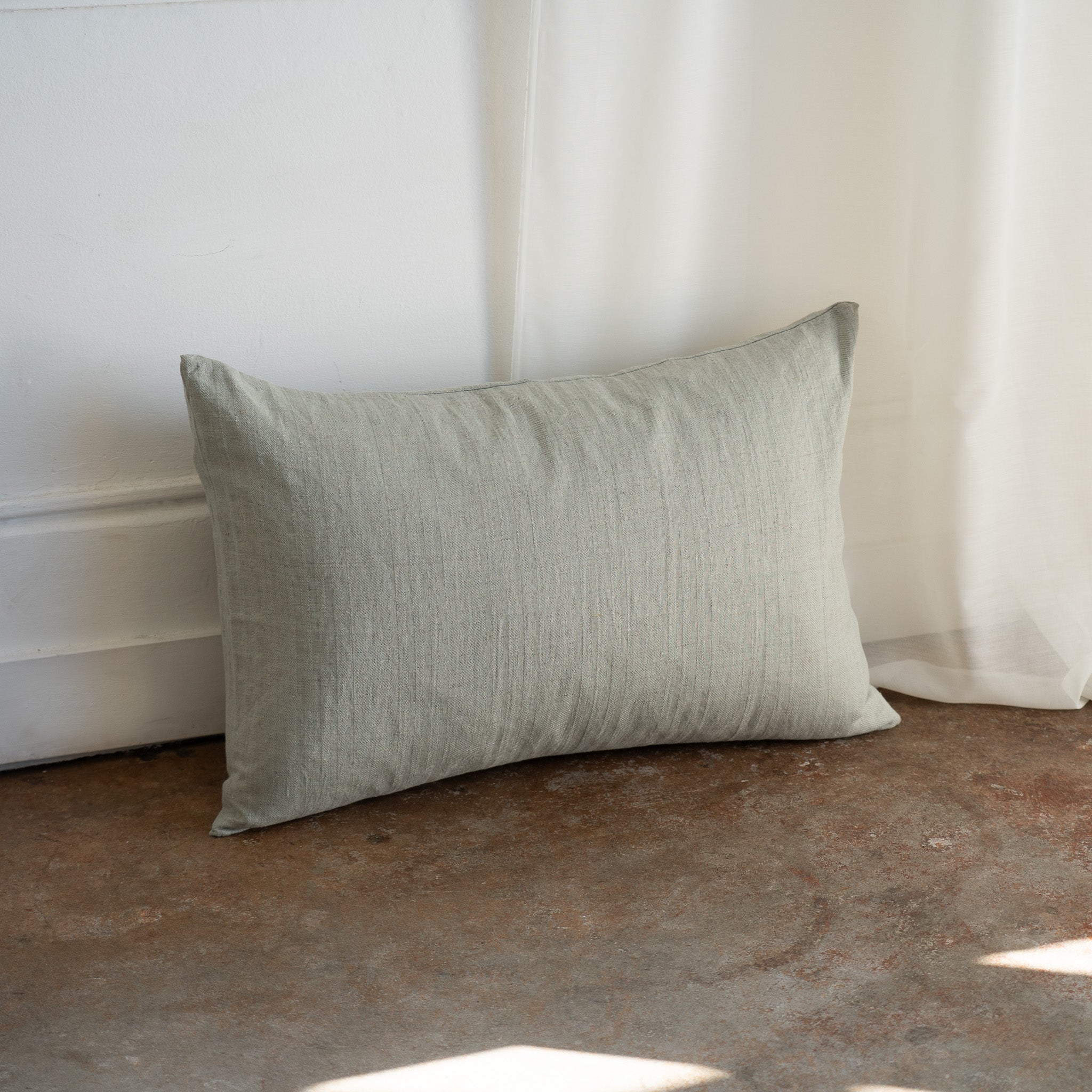 A rectangular linen throw pillow in a muted sage green color, leaning against a white wall on a textured brown floor with soft natural light filtering in from the right.