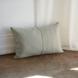 A rectangular linen throw pillow in a muted sage green color, leaning against a white wall on a rustic concrete floor with a white curtain in the background.