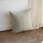 A square linen throw pillow in a light sage green color, leaning against a white wall on a rustic concrete floor next to a sheer curtain.