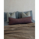 A styled bed with a textured taupe linen duvet, a dark red patterned lumbar pillow, and slate blue pillows against a textured off-white wall.