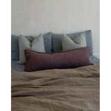 A styled bed with a textured taupe linen duvet, a dark red patterned lumbar pillow, and slate blue pillows against a textured off-white wall.