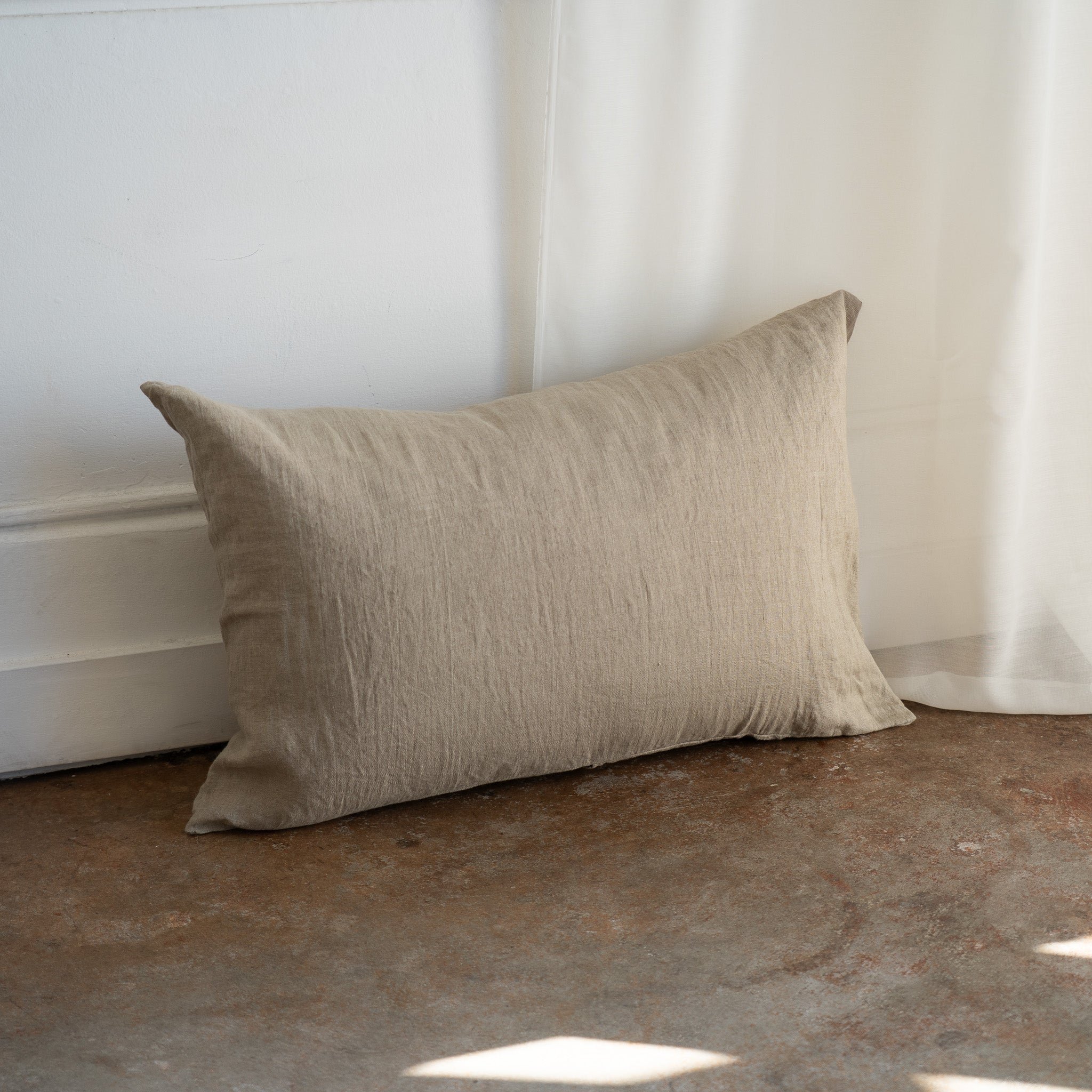 A rectangular beige linen throw pillow with a textured crinkled fabric, leaning against a white wall on a rustic concrete floor with soft natural light filtering in from the right.