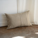 A rectangular beige linen throw pillow with a central vertical seam, leaning against a white wall on a rustic concrete floor next to a white curtain.
