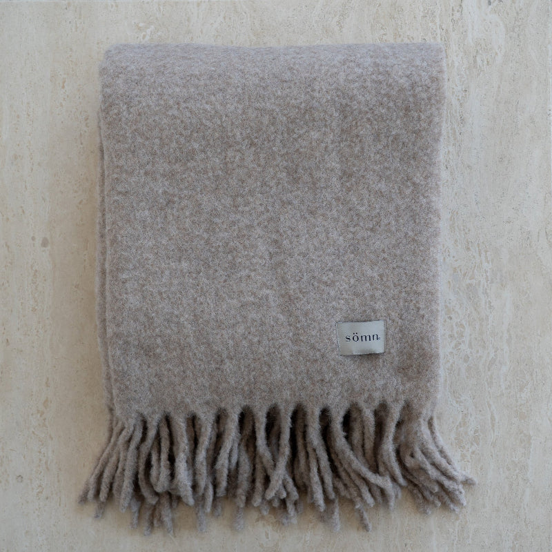 Gray blanket with fringes and a visible brand label on a textured beige background