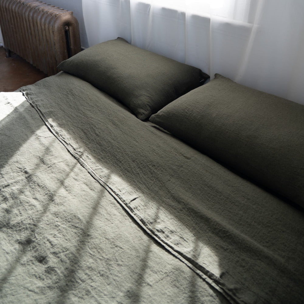 Bed with olive green bedding and pillows in a room with a radiator and window.