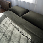 Bed with olive green bedding and pillows in a room with a radiator and window.