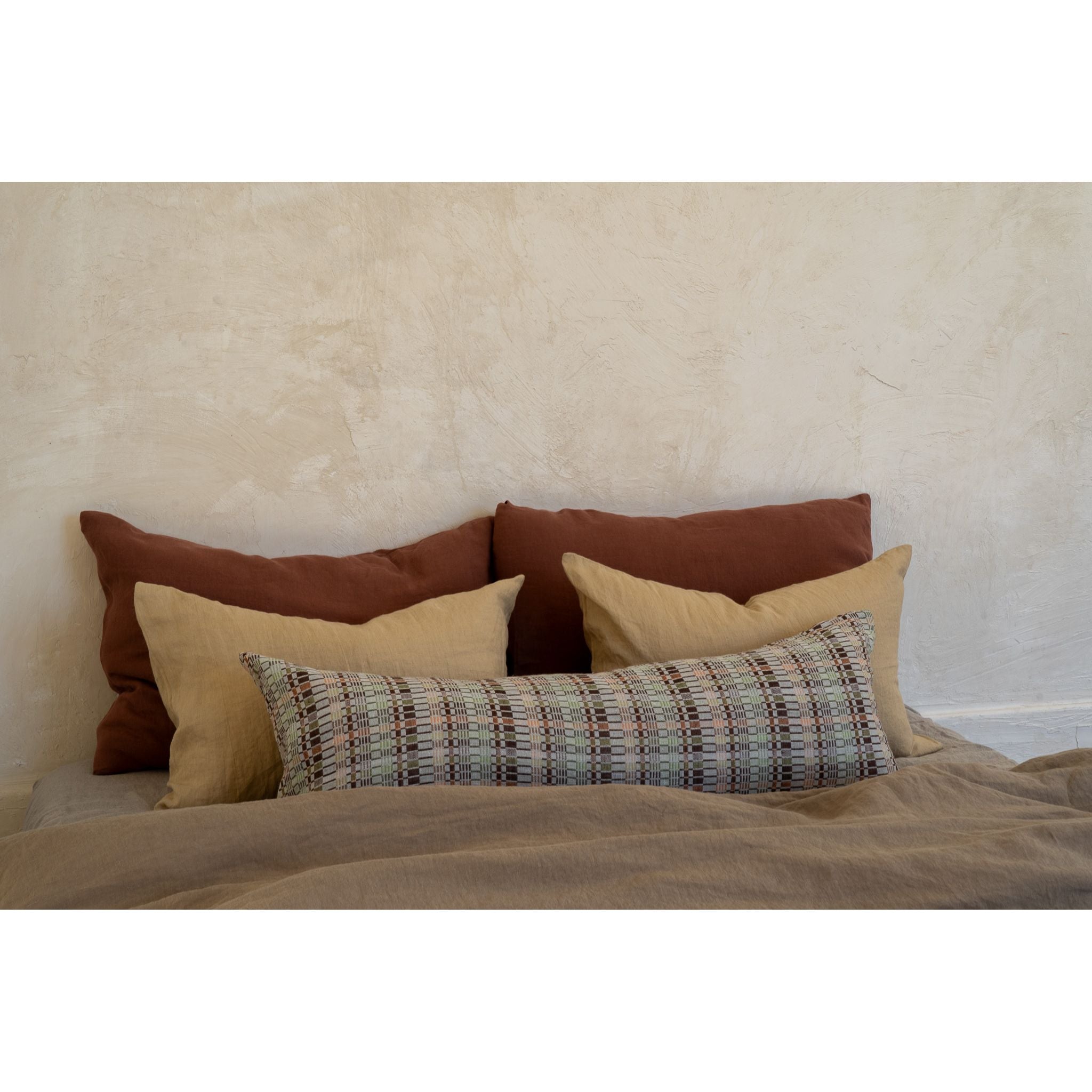 A styled bed setup featuring a collection of textured throw pillows in warm earthy tones, including rust brown, beige, and a plaid patterned lumbar pillow, set against a rustic plaster wall.