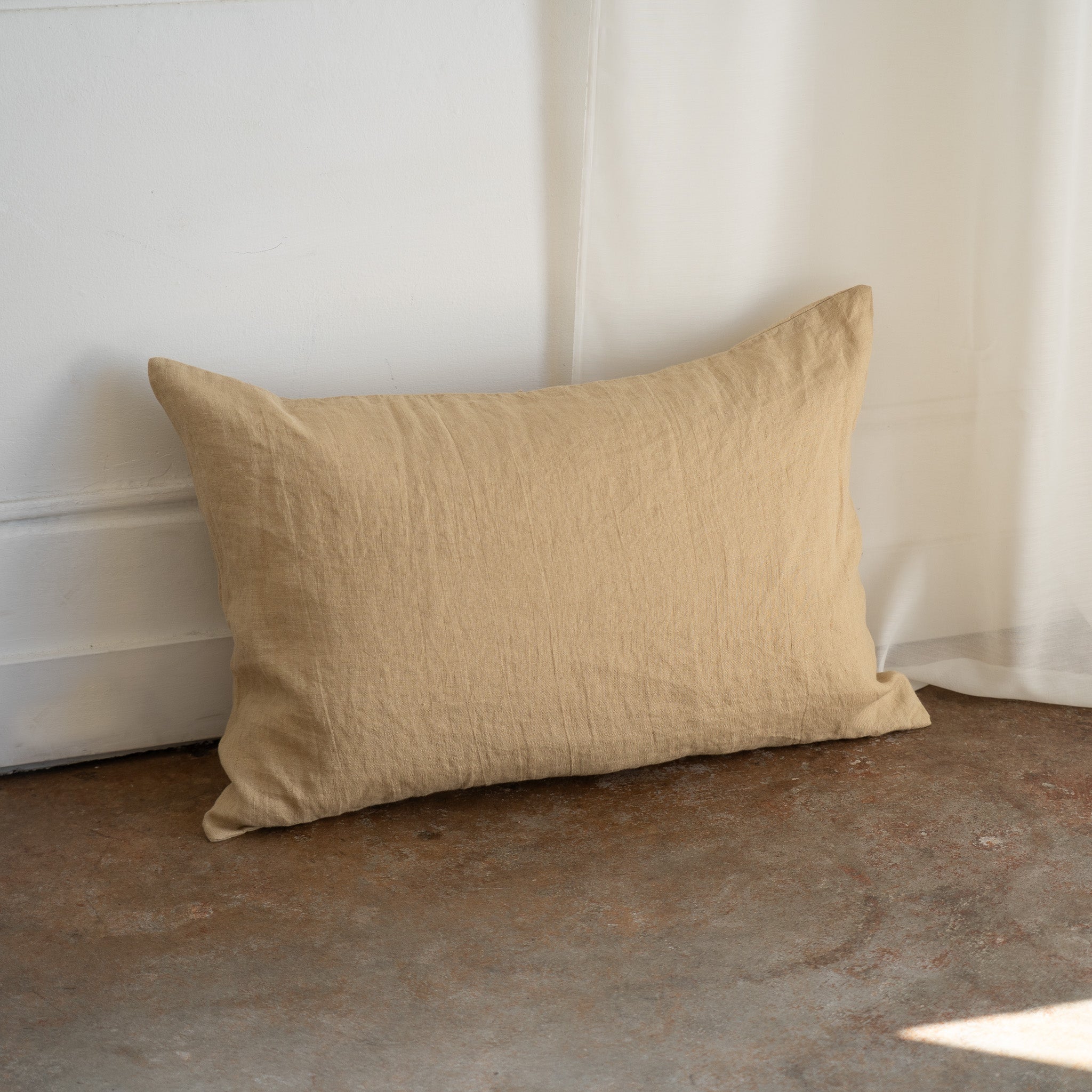 A rectangular beige linen throw pillow with a textured, crinkled fabric, leaning against a white wall on a rustic concrete floor.