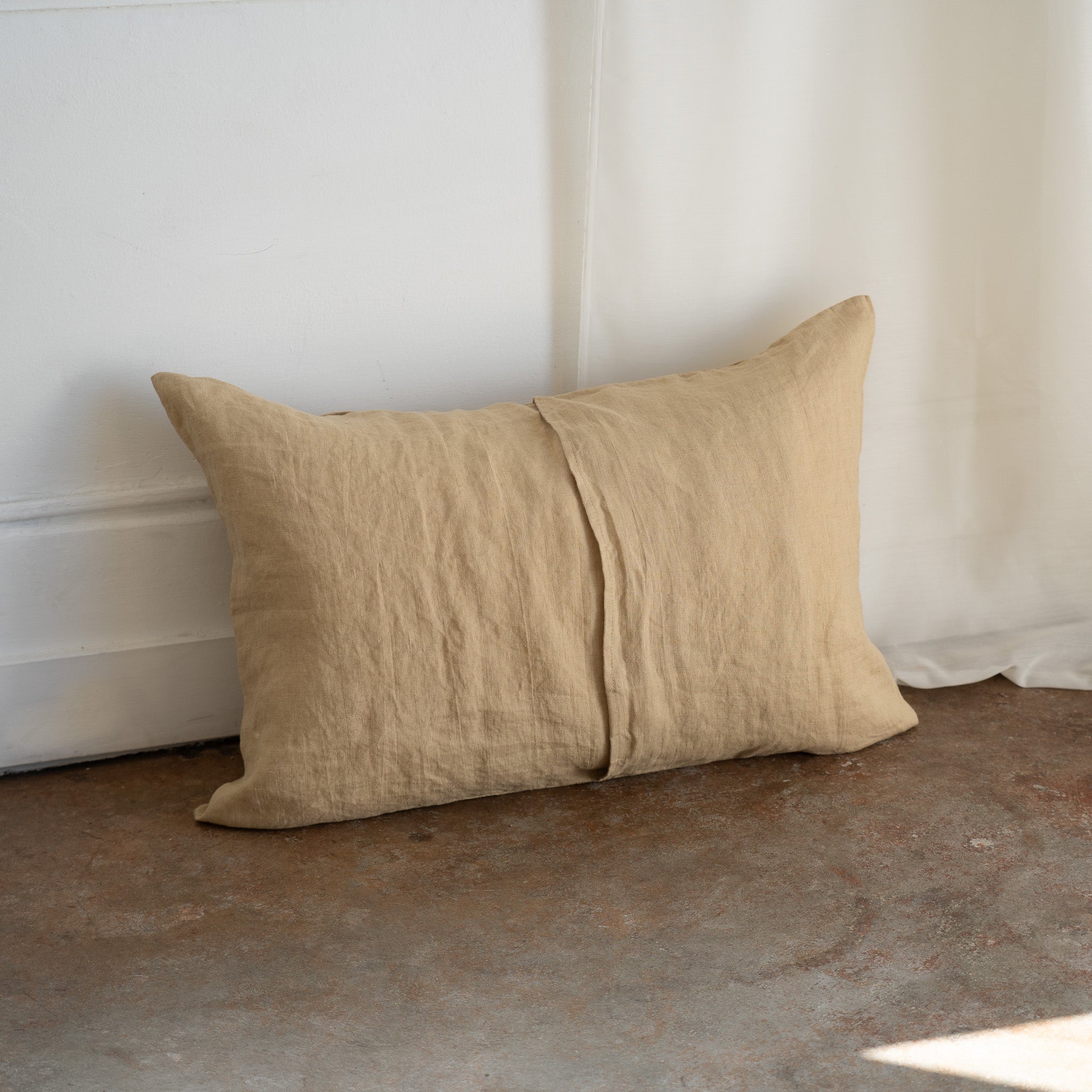 A rectangular beige linen throw pillow with a central seam, leaning against a white wall on a textured brown floor.