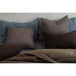 A styled collection of home textiles featuring brown and beige patterned pillows, solid dark brown pillows, and a matching brown throw blanket draped over a bed with slate blue pillows and a textured off-white wall behind.